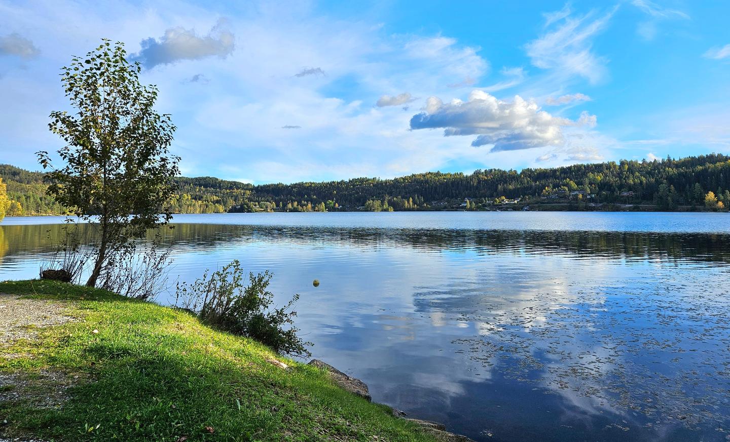 Hagatjern i Mjøndalen en høstdag, med blank vannflate, skogkledde åser og blå himmel.